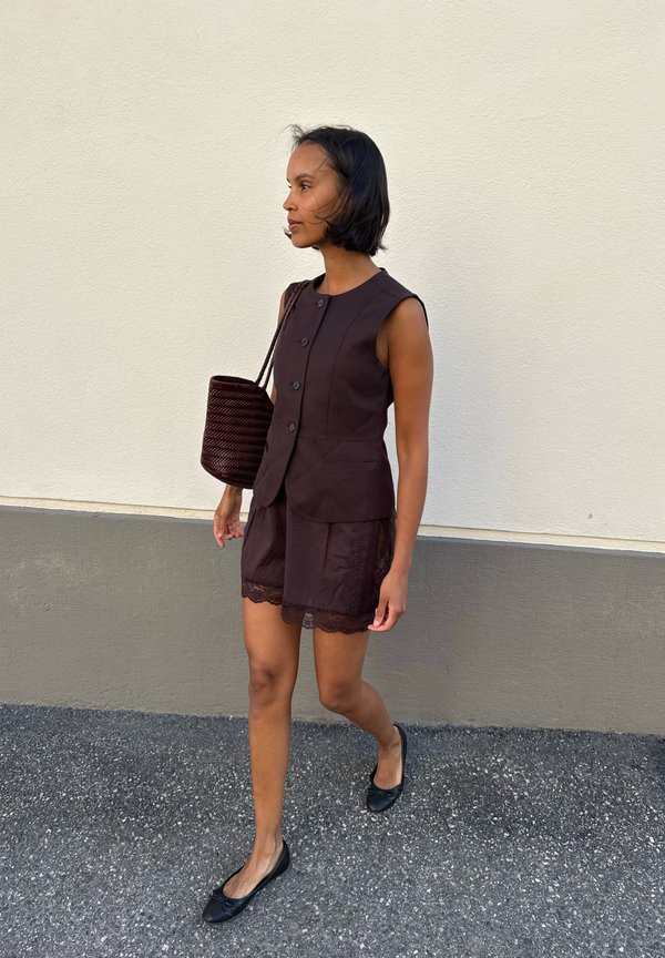 Mujer caminando al aire libre con una blusa marrón sin mangas con botones, pantalones cortos a juego con borde de encaje, bailarinas negras y llevando un bolso marrón tejido al hombro.