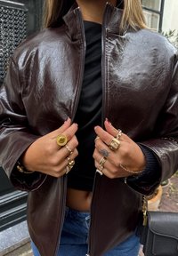 Hnedá bunda z umelého koženého materiálu s vysokým golierom, zapínaním na zips a spustenými ramenami. Nosená cez čierne tričko, doplnená viacerými prsteňmi.