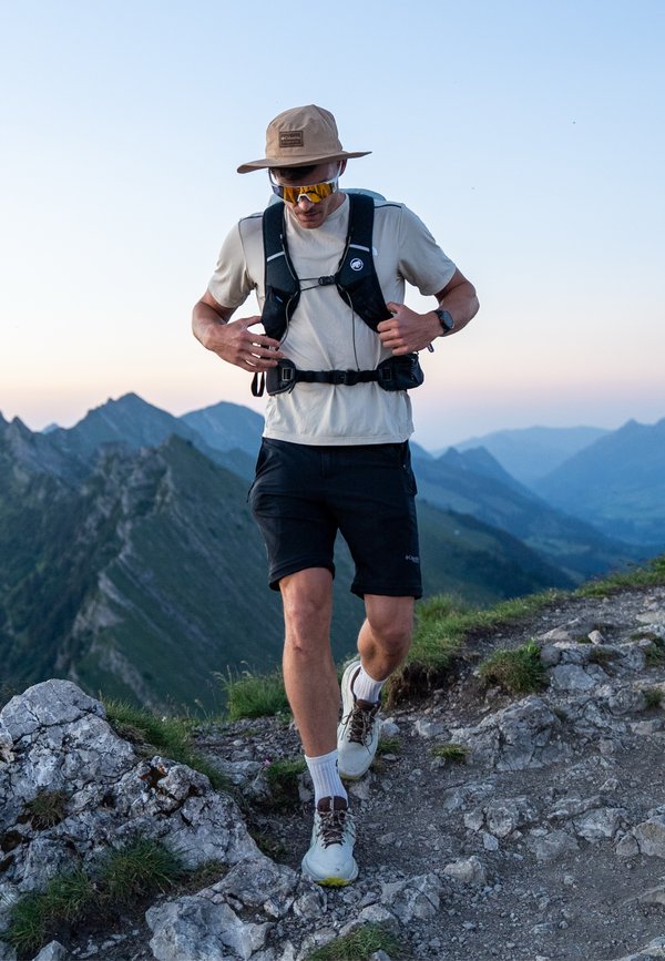 Homme portant un chapeau, des lunettes de soleil et un équipement de randonnée, marchant sur un sentier montagneux rocheux tôt le matin ou tard le soir.