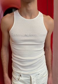 Personne portant un débardeur blanc côtelé et un pantalon, avec un texte discret "Ludovic de Saint Sornin" sur la poitrine, posant devant un fond rouge.