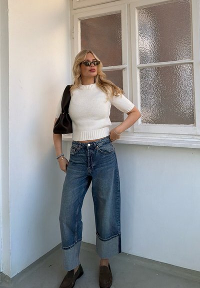 Mujer con jersey blanco de manga corta, vaqueros de pierna ancha con puños, gafas de sol negras, bolso negro al hombro y mocasines oscuros, de pie junto a una ventana de vidrio texturizado.