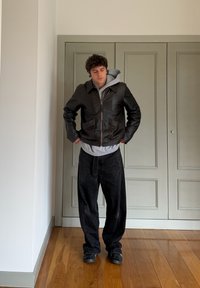 Jeune homme debout à l'intérieur, portant une veste en cuir noire par-dessus un sweat à capuche gris, un pantalon noir ample et des baskets noires sur un sol en bois.