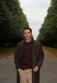 Modell i en brun kappa över en vinröd tröja och beige byxor, som går längs en trädkantad stig med höstlöv.