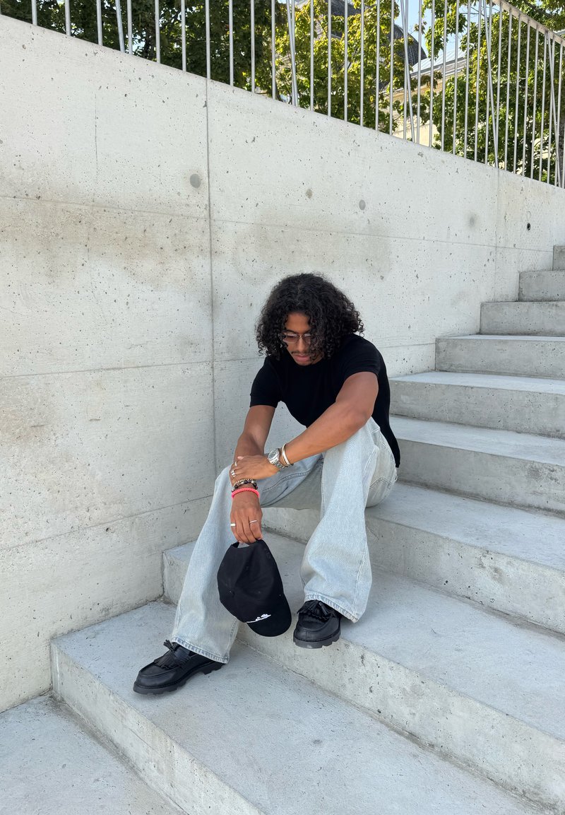 T-shirt noir, jeans bleu clair et chaussures noires. Tenant une casquette noire. Assis sur des escaliers en béton avec un mur texturé en arrière-plan.