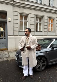 Beige ullfrakk, kremfarget genservest, hvite bukser, brune lærhansker, holder en kaffekopp. Parkert grå Audi-bil i bakgrunnen.