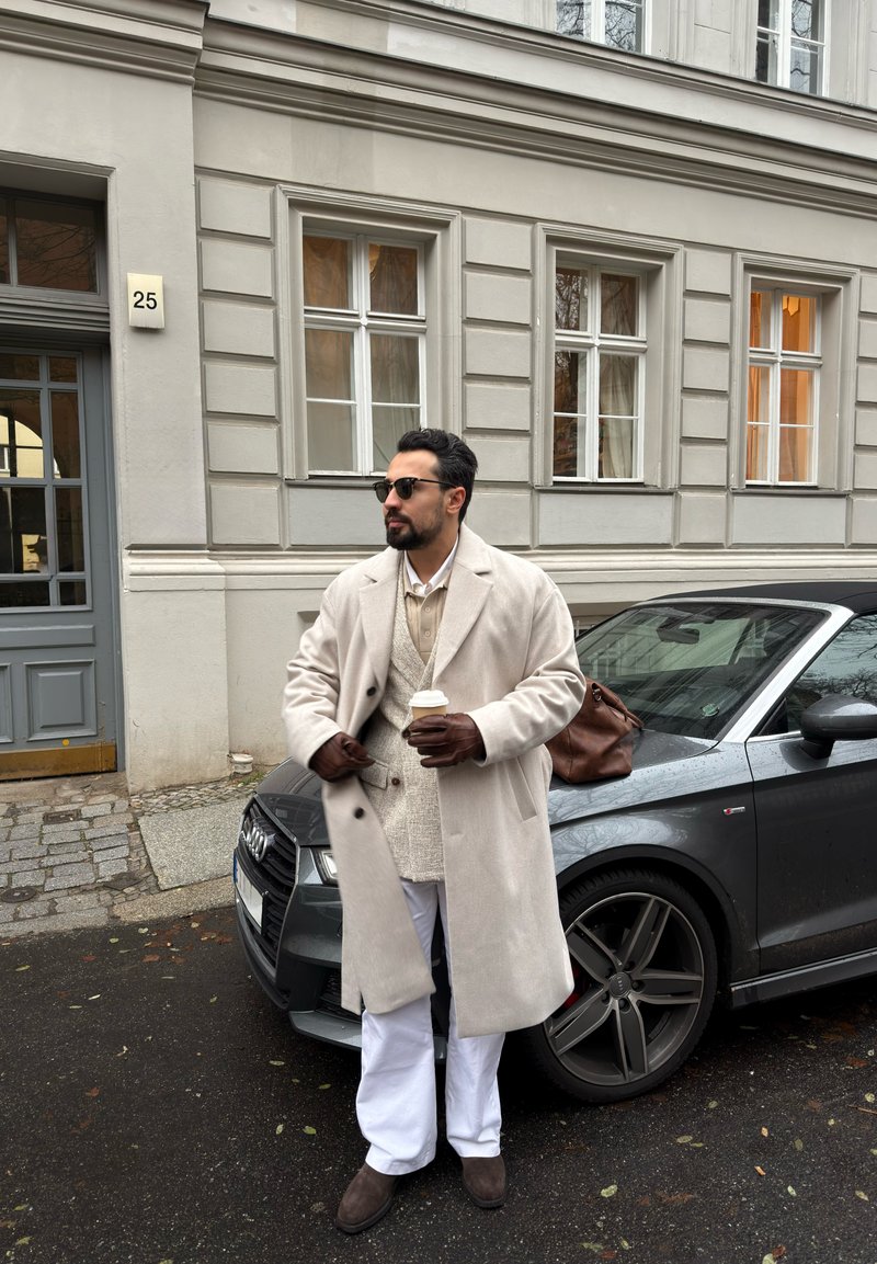 Beige wollen overjas, crèmekleurig sweatervest, witte broek, bruine leren handschoenen, met een koffiekopje in de hand. Parkeergrijze Audi-auto op de achtergrond.