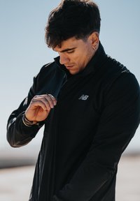 Czarna kurtka na zamek z wysokim kołnierzem, z małym logo New Balance na lewej piersi. Materiał wydaje się gładki i lekki.