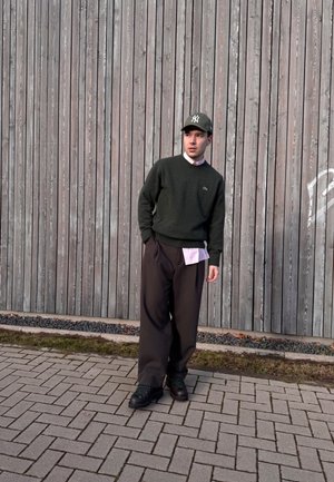 Suéter verde oscuro, camisa de cuello rosa claro debajo, pantalones negros holgados, zapatillas verdes y una gorra ajustada oscura, de pie contra una pared de madera.