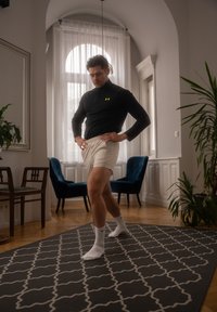 Haut de sport zippé noir avec logo néon, shorts beiges, chaussettes blanches, debout sur un tapis gris à motifs dans une pièce bien éclairée avec des plantes.