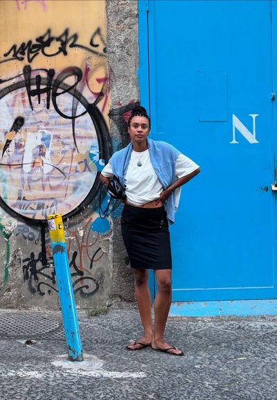 Una persona con una camisa blanca y una falda negra está de pie frente a una pared cubierto de graffiti, sosteniendo un casco. Se pueden ver una puerta azul y un poste amarillo.