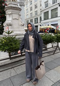Veste gris mélange laine à silhouette oversize, pull en maille beige, pantalon gris à jambes larges et sac fourre-tout à motifs. Cadre urbain avec végétation.