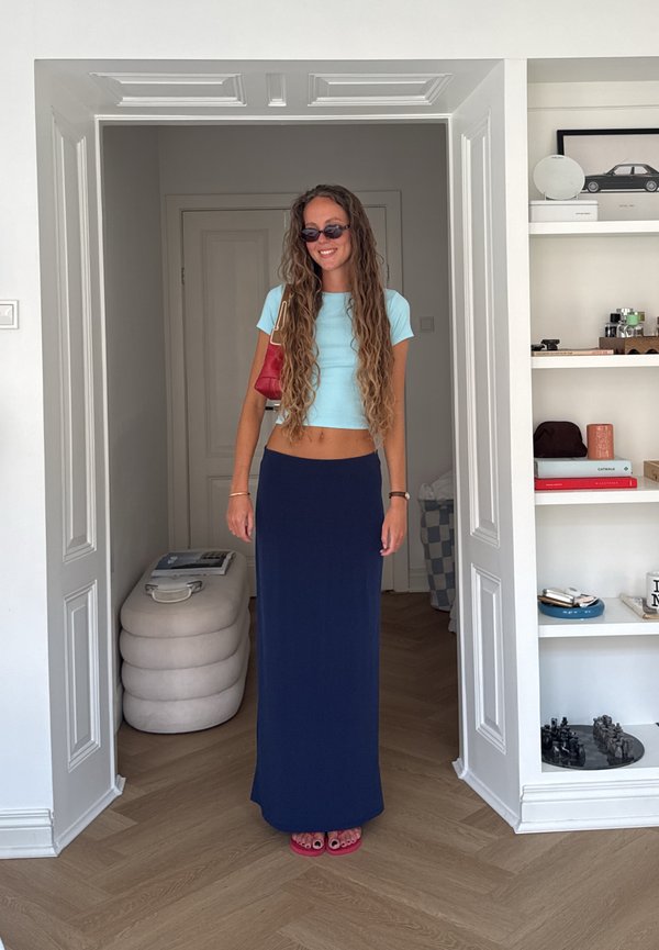 Jonge vrouw met lang krullend haar, die een zonnebril, een lichtblauw cropped topje, een donkerblauwe lange rok en rode sandalen draagt en binnen staat bij witte planken.