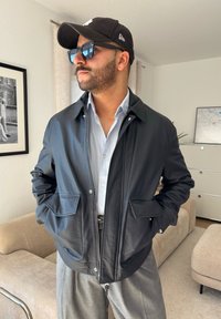 Casaco de cabedal preto com fecho à frente, dois bolsos frontais e colarinho, combinado com uma camisa azul clara de botões e cal�ças cinzentas.