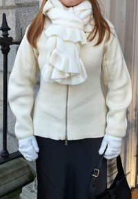 Personne portant une écharpe blanche à volants, un pull à fermeture éclair crème, des gants blancs, et tenant un sac à main noir devant un mur en pierre et une rambarde.