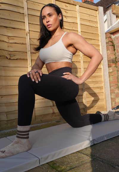 Mujer con top deportivo y leggings estira sobre una esterilla gris al aire libre, haciendo una zancada con una rodilla doblada y las manos apoyadas en el muslo.