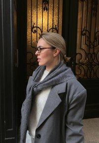 Veste grise oversize avec de grands revers, portée sur un pull léger. Écharpe grise douce drapée autour du cou. Lunettes écaille de tortue marron.