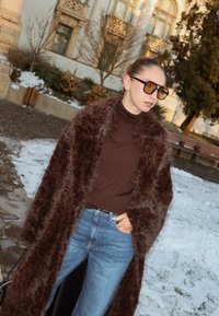 Femme portant un manteau en fausse fourrure marron oversize, un col roulé marron, un jean bleu et des lunettes de soleil teintées, debout à l'extérieur près d'un sol recouvert de neige.