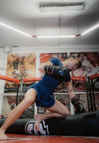 Naine sinises spordirõivastuses ja poksikinnastega põlvitamas musta poksikoti peal jõusaalis, kus on seinapildid ja traataed.