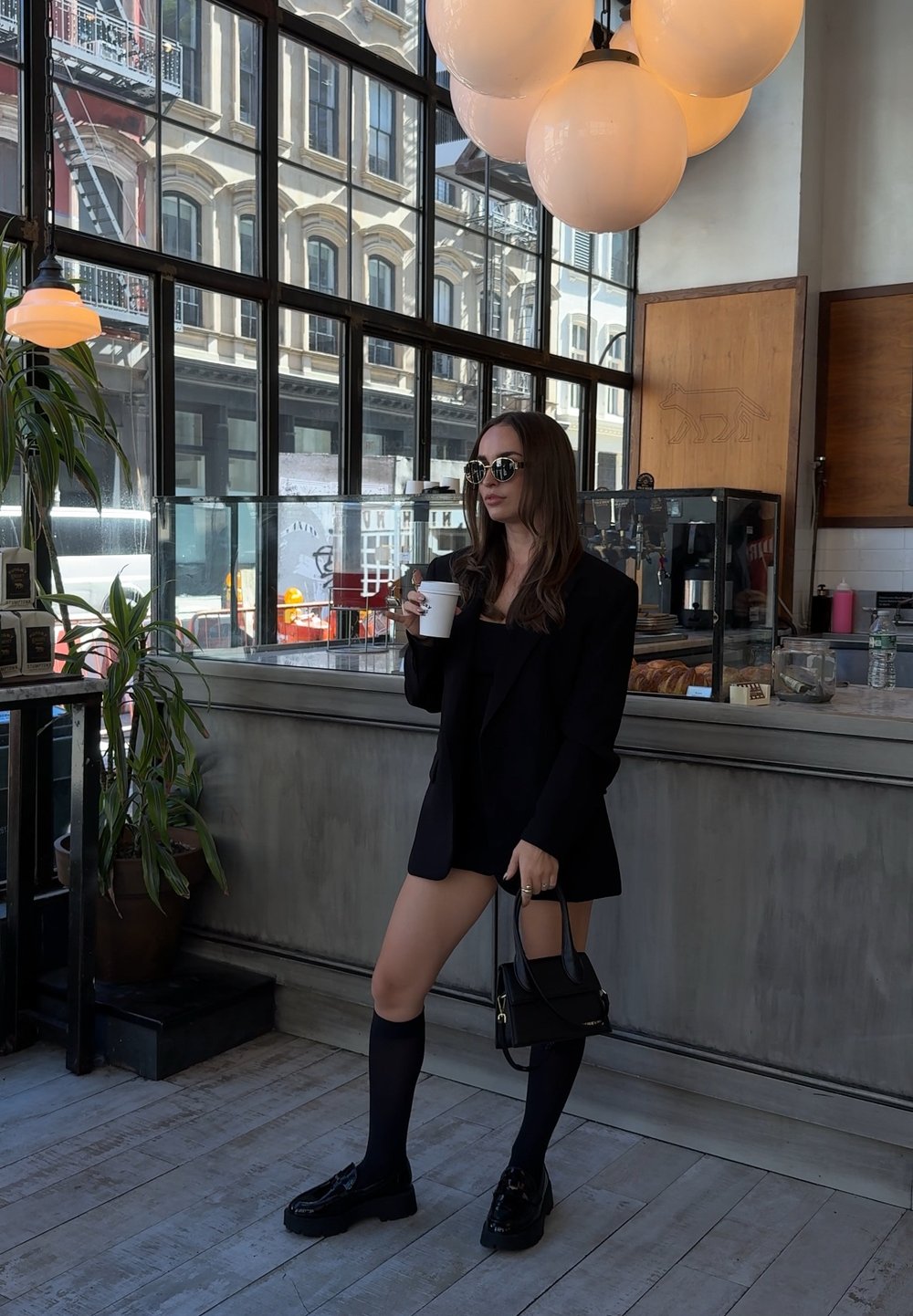Črn oversized blazer, prilegajoča črna majica in hlače. Črne nogavice do kolen z sijajnimi loafri. Drži belo papirnato skodelico, stoji v kavarni.