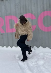 Personne portant une veste beige en peluche, une casquette noire, un pantalon sombre et des bottes, debout sur un sol enneigé, levant un pied, devant un mur gris avec de grandes lettres roses.