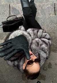 Szary futrzany płaszcz z szerokimi rękawami, czarne skórzane rękawiczki i wąskie czarne spodnie. Czarna torebka spoczywa na teksturowanej szarej powierzchni.