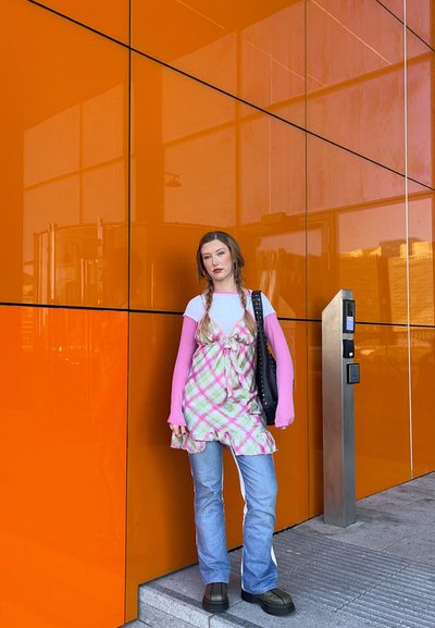 Joven mujer con el cabello trenzado que lleva un vestido rosa con capas y de cuadros sobre vaqueros, de pie junto a una pared reflectante naranja al lado de un terminal de pago metálico.