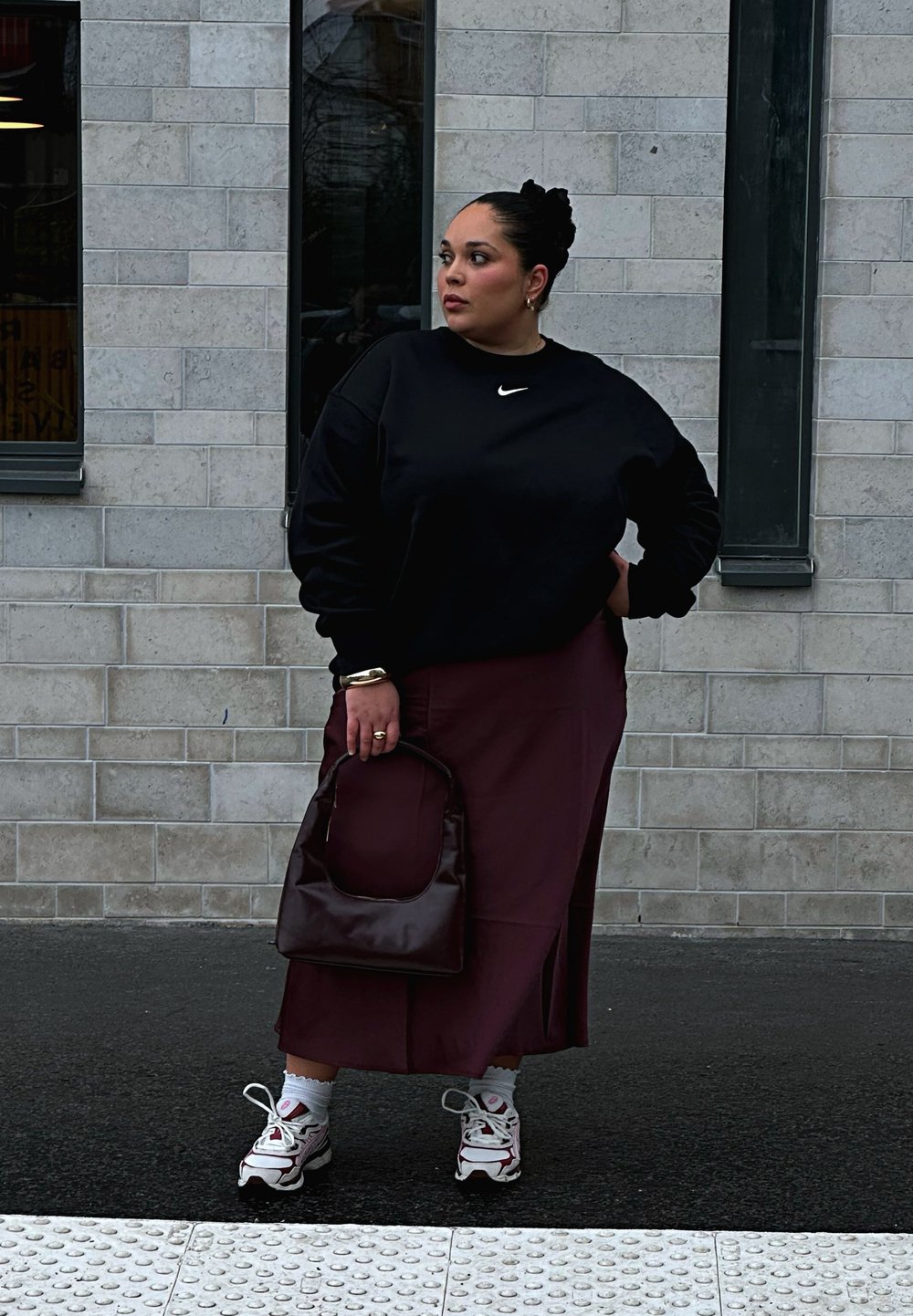 Czarna bluza Nike, burgundowa spódnica z bocznym rozcięciem, brązowa torebka, białe skarpetki i wielokolorowe trampki. Tło ze strukturalną ścianą.
