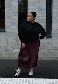Czarna bluza Nike, burgundowa spódnica z bocznym rozcięciem, brązowa torebka, białe skarpetki i wielokolorowe trampki. Tło ze strukturalną ścianą.