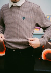 Långärmad pikétröja i ett brunt och vitt rutmönster, med en vit krage och en grön logotyp. Bärs över svarta manchesterbyxor.