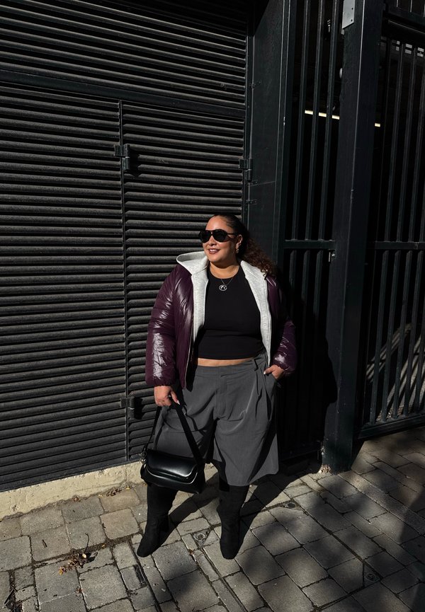 Una mujer con gafas de sol y una chaqueta color ciruela posa en una acera de la ciudad, sosteniendo un bolso negro junto a una persiana metálica oscura.