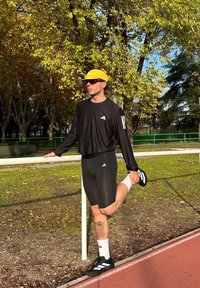 Camisa negra de manga larga, pantalones cortos negros, gorra amarilla y gafas de sol. De pie en una pista, estirando una pierna contra una barandilla blanca.