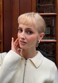 Cárdigan blanco de punto con cuello y cremallera, llevado por una persona de cabello rubio claro, maquillaje suave y pendientes de aro plateados.