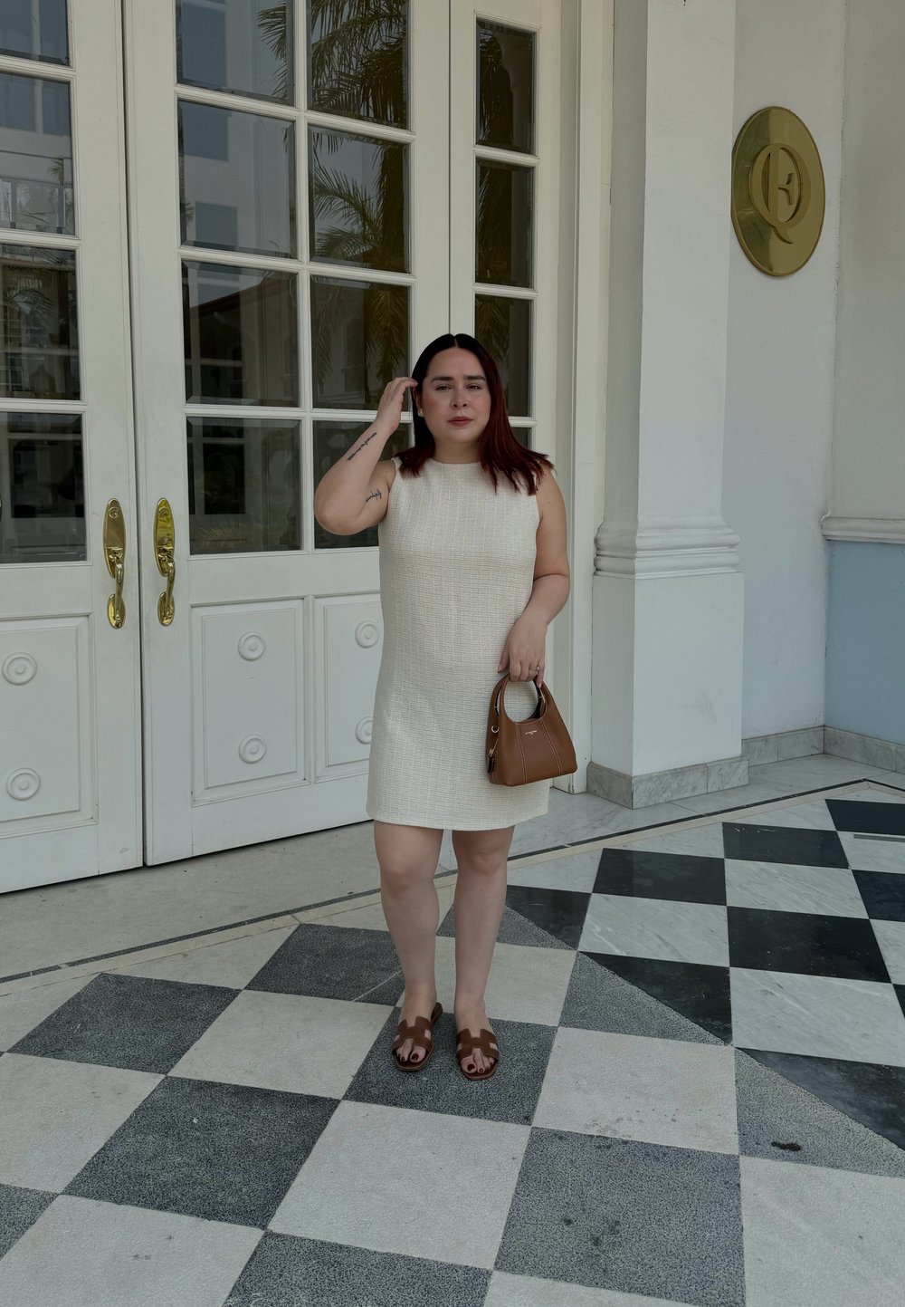 Robe sans manches blanche et texturée avec une coupe droite, assortie à des sandales marron et un petit sac à main marron. Debout sur un sol à motifs noir et blanc.