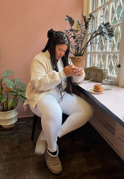 Modelo vistiendo una chaqueta bomber de felpa color crema y leggings blancos, sentada a una mesa con un croissant y un bolso verde, rodeada de plantas.