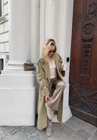 Femme en manteau long beige ajustant son pantalon tout en tenant une tasse de café, debout près d'une grande colonne blanche et d'une porte en bois ornée avec un sac à main posé sur le rebord.