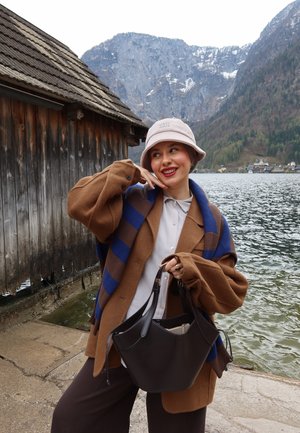 Joven mujer con abrigo marrón y sombrero beige sonriendo junto a una cabaña de madera junto al lago con montañas y agua al fondo.