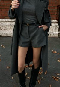 Un manteau gris foncé sur un haut gris assorti et un short ajusté, avec une ceinture noire arborant un logo doré, associé à des bottes noires montant jusqu'aux genoux.