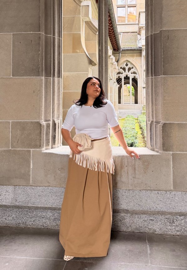 Vrouw in wit topje en beige rok met een clutch, staand in een stenen boog met een gotische stijl gebouw en tuin op de achtergrond.