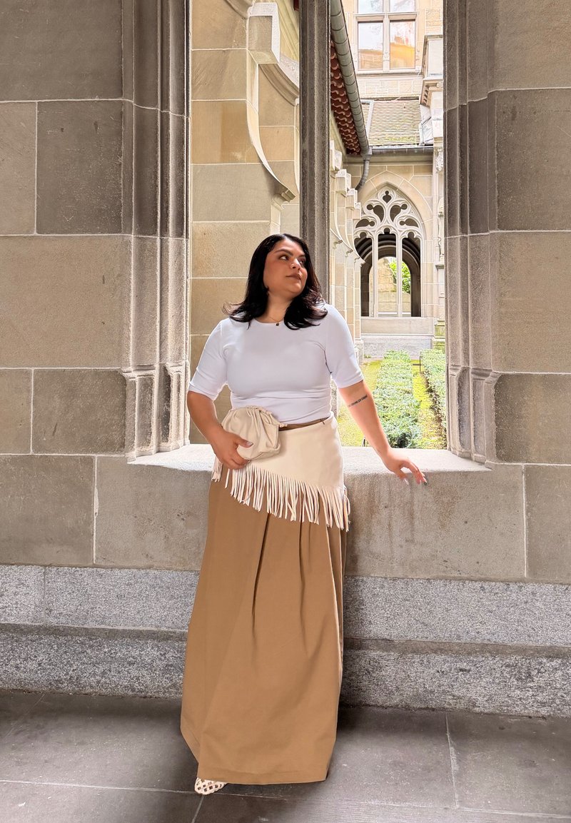Frau in weißem Top und beiger Rock hält eine Clutch, steht in einem steinernen Torbogen mit gotischem Gebäude und Garten im Hintergrund.