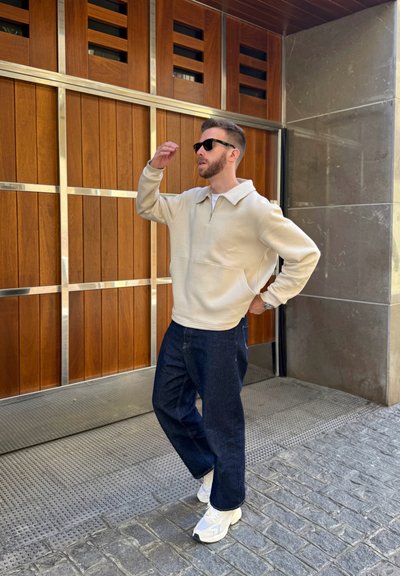 Sudadera beige con cremallera y cuello, combinada con unos vaqueros oscuros de corte relajado y zapatillas blancas. El fondo cuenta con paneles de madera.
