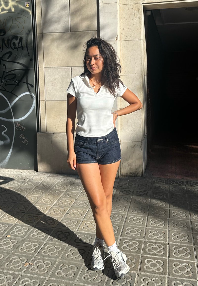Polo shirt bianco a coste, pantaloni corti di jeans scuri, calzini bianchi e sneakers grigio chiaro. In piedi alla luce del sole con un'ombra su un pavimento texturizzato.