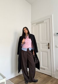Gelaagde outfit met een roze gebreid vest, een gestreept hemd en wijde bruine broek, aangevuld met een zwarte overjas en donkere schoenen.