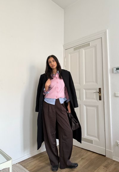 Conjunto superpuesto que incluye un cárdigan de punto rosa, camisa de rayas y pantalones anchos marrones, complementado con un abrigo negro y zapatos oscuros.
