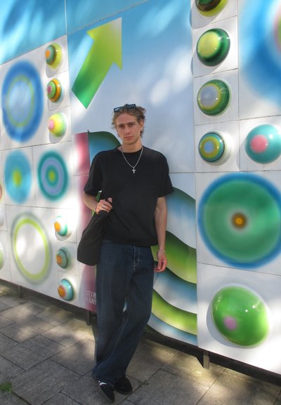 Camiseta negra y jeans oscuros llevados por una persona apoyada contra una pared colorida con diseños circulares en patrones verdes, azules y rosas.