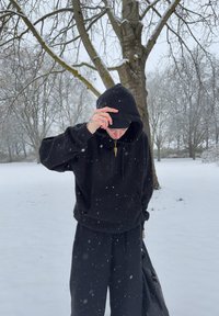 Sort hettegenser med glidelås i halsen, kombinert med matchende svarte joggebukser, stående i fallende snø med nakne trær i bakgrunnen.