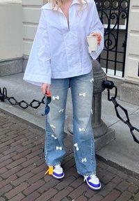 Camisa blanca de gran tamaño con un profundo cuello en pico, jeans de color azul claro adornados con aplicaciones de moño blanco y zapatillas con acentos morados.