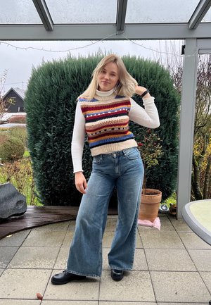 Chaleco de mezcla de lana con rayas horizontales de colores, combinado con jeans de denim de pierna ancha de tiro alto. Escenario al aire libre con vegetación al fondo.