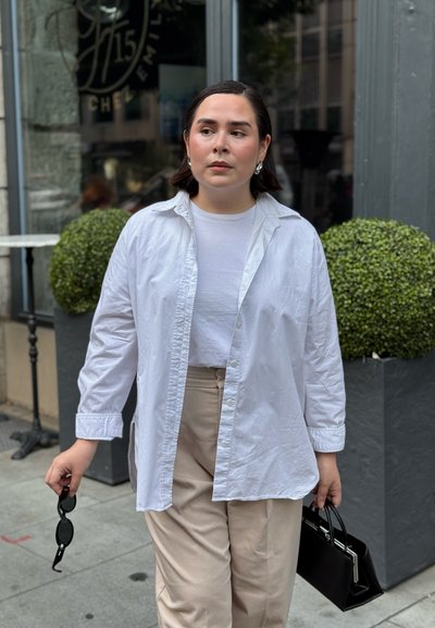 Adulto vestido con camisa blanca, pantalones beige, sosteniendo gafas de sol y un bolso negro, caminando por la acera urbana cerca de una tienda y plantas decorativas.
