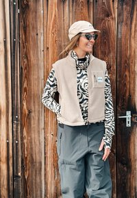 Femme portant une casquette beige, des lunettes de soleil, un gilet polaire beige, une chemise à manches longues à motifs et un pantalon gris, debout devant une porte en bois.