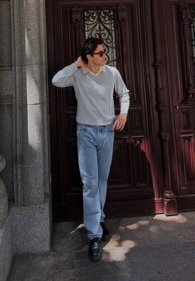 Joven con gafas de sol, suéter ligero y vaqueros se apoya en una gran puerta de madera oscura bajo el sol cerca de una pared de piedra.